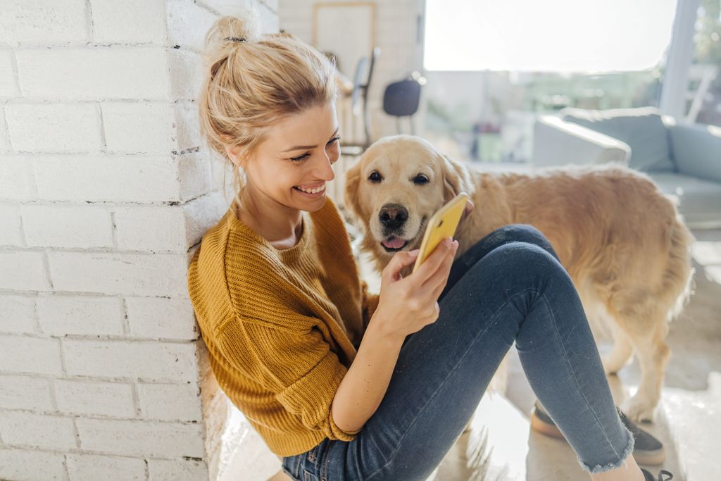 Woman is sitting on the floor with mobile device playing and her dog sitting next to her.